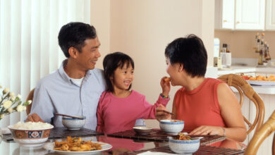 An Asian Family, An Adult Male And Female Are Seated Around A Table Eating A Meal With A Young Female Standing In Between The Adults.